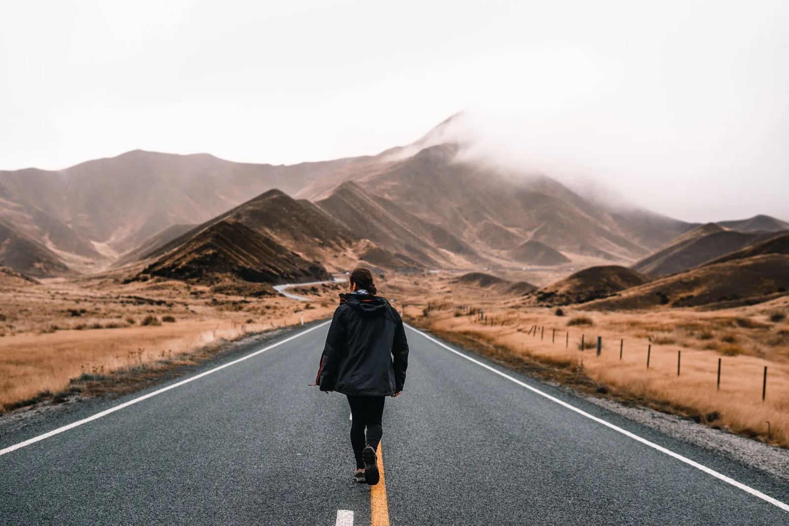 A walker in a dark jacket on an empty two-lane road through brown hills under low cloud