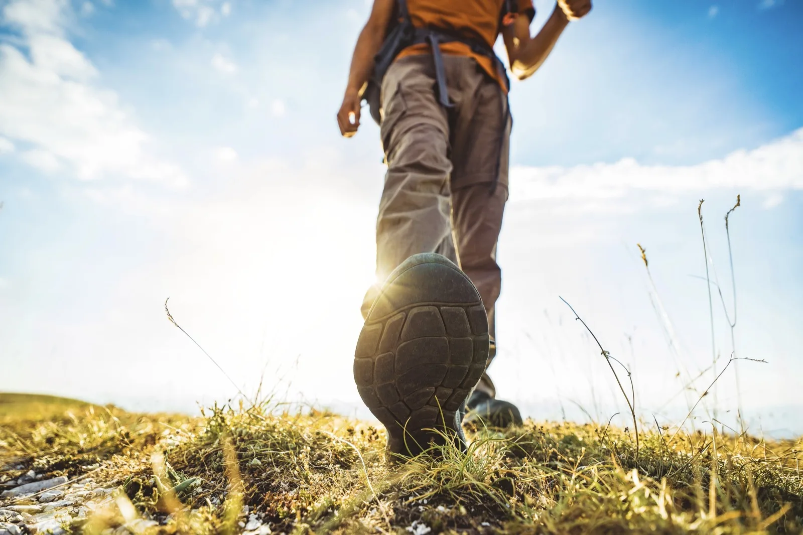 A hiker's boot mid-stride on a grassy hilltop path, seen from ground level against the sun